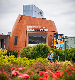 Exterior view of the College Football Hall of Fame building with people walking nearby and colorful foliage in the foreground.