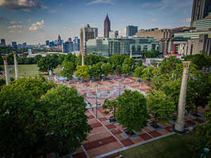 Aerial view of a city park with green trees, a patterned brick plaza, and tall buildings in the background under a partly cloudy sky.