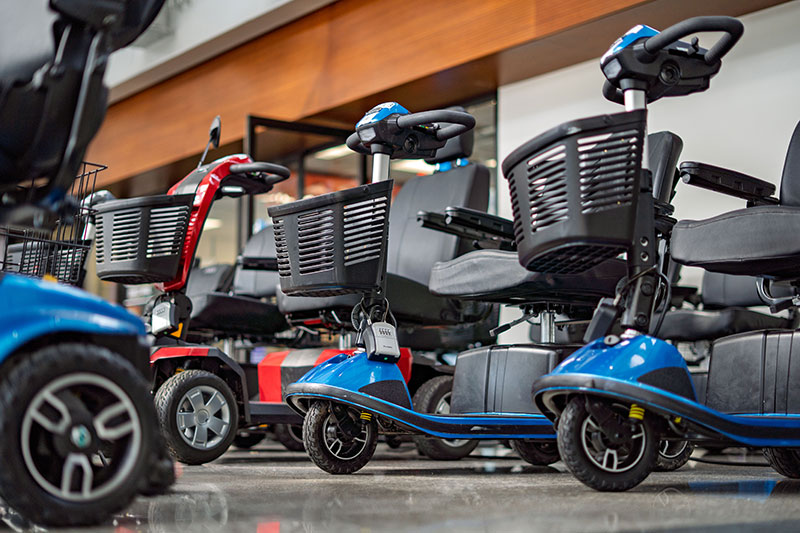 Row of mobility scooters with black seats and baskets, featuring blue and red models, displayed indoors on a shiny floor.