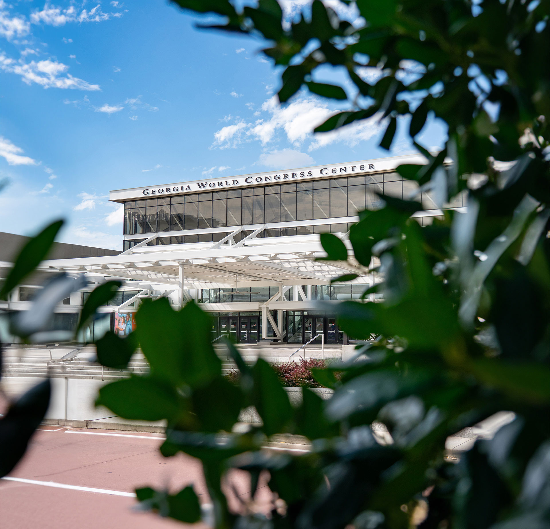 Exterior view of the Georgia World Congress Center building framed by green leaves under a partly cloudy blue sky.