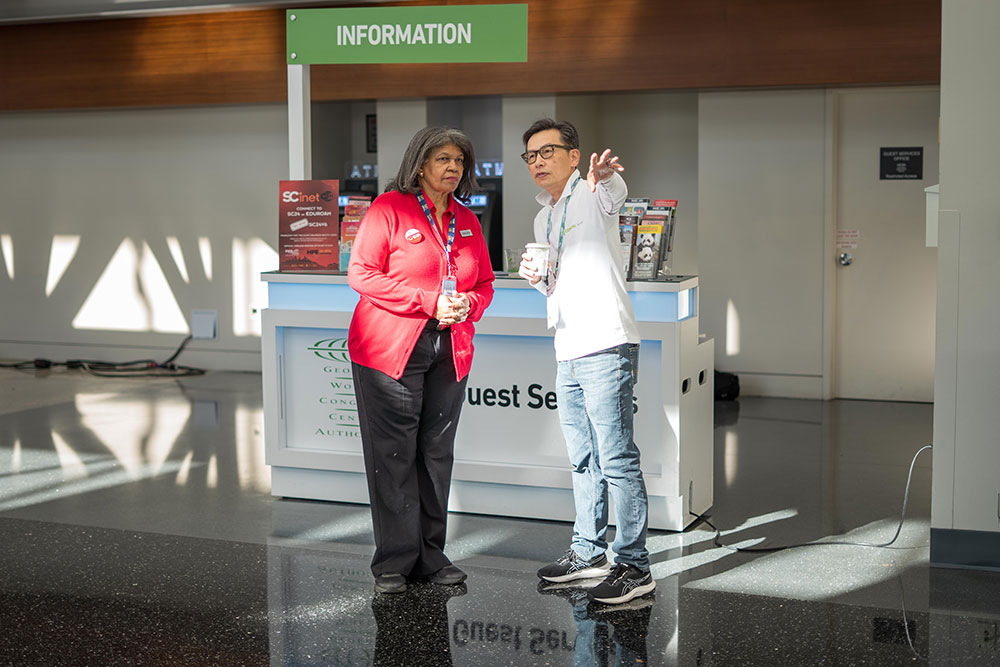 Two people standing and talking in front of an information desk; one person wearing a red jacket and the other pointing while holding a cup.