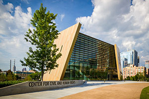 Modern angular building of the Center for Civil and Human Rights under a partly cloudy sky with a tree in front.