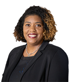 Portrait of a smiling woman with curly hair wearing a black blazer and silver necklace in an indoor setting.